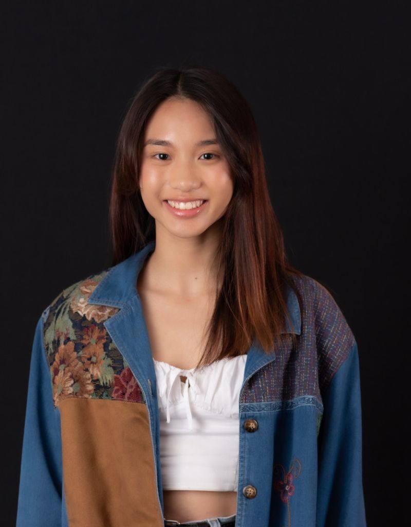 A young Asian woman, shoulder-length dark brown wearing a white shirt and a blue-brown jacket on top, smiling at the camera