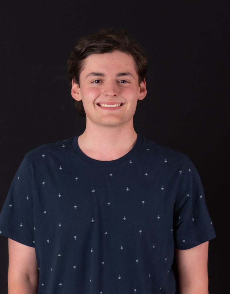 A young man, short hair wearing a navy blue, polka dotted, short sleeve t-shirt smiling at the camera.