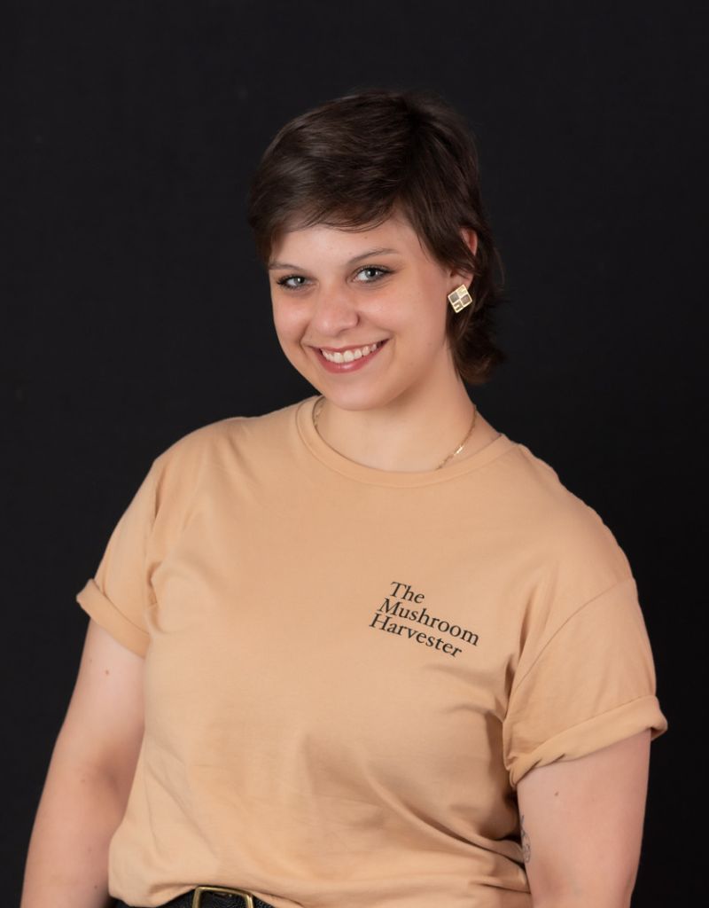 A young person, short hair, wearing a short sleeve cream t-shirt, smiling at the camera