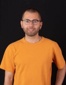 A young man, with a slight bear and hear, Black framed eyeglasses, wearing an orange short sleeve t-shirt, smiling at the camera.