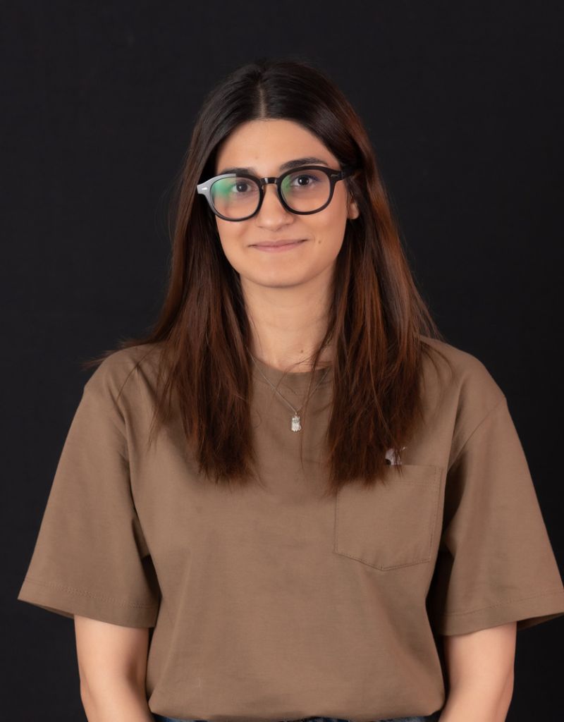 A young woman with an armpit length brown hair, black framed Square eyeglasses, wears a light brown short sleeve t-shirt, smiles at the camera