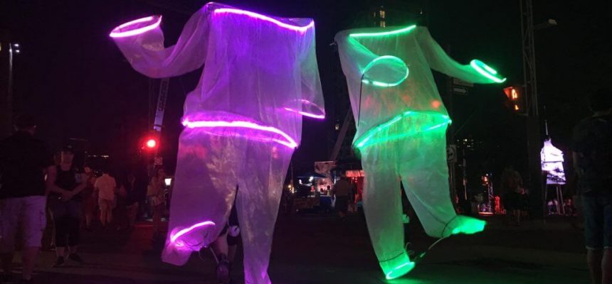 Two Giant puppets dancing with people while bright LED strips border their head, arms and feet.