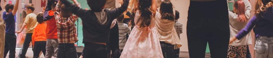 anna-earl-XBDHmIXvsvM-unsplash Children and parents jumping and clapping their hands above their head during a creative dance workshop.