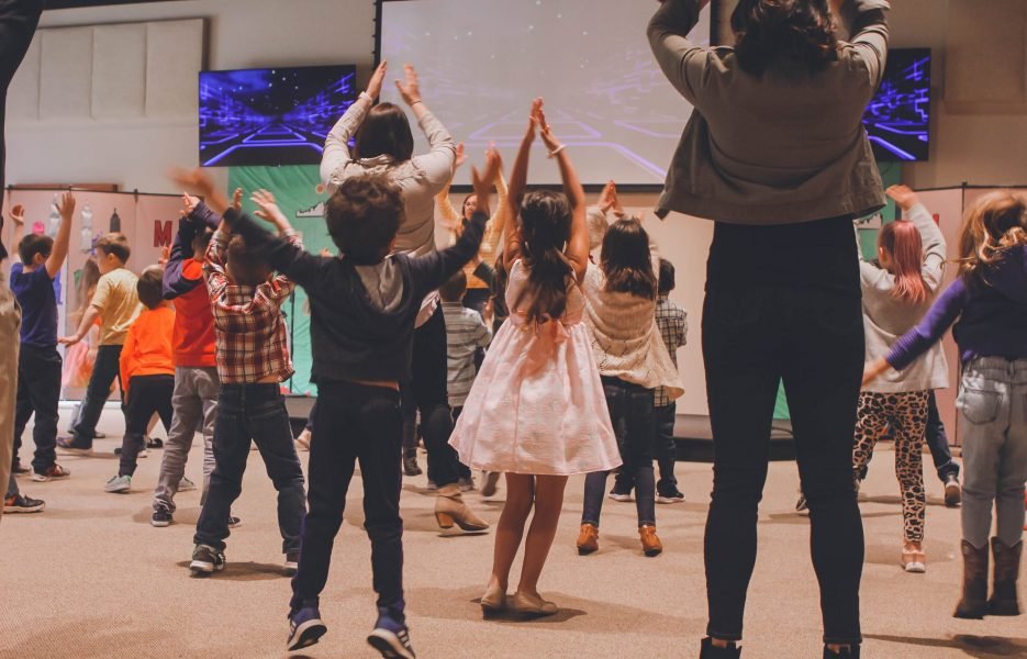 anna-earl-XBDHmIXvsvM-unsplash Children and parents jumping and clapping their hands above their head during a creative dance workshop.