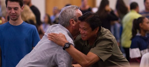 erika-giraud-7KhbREQPFD4-unsplash An elderly man hugs another man in a community gathering with people of several walks of life socializing in the background.